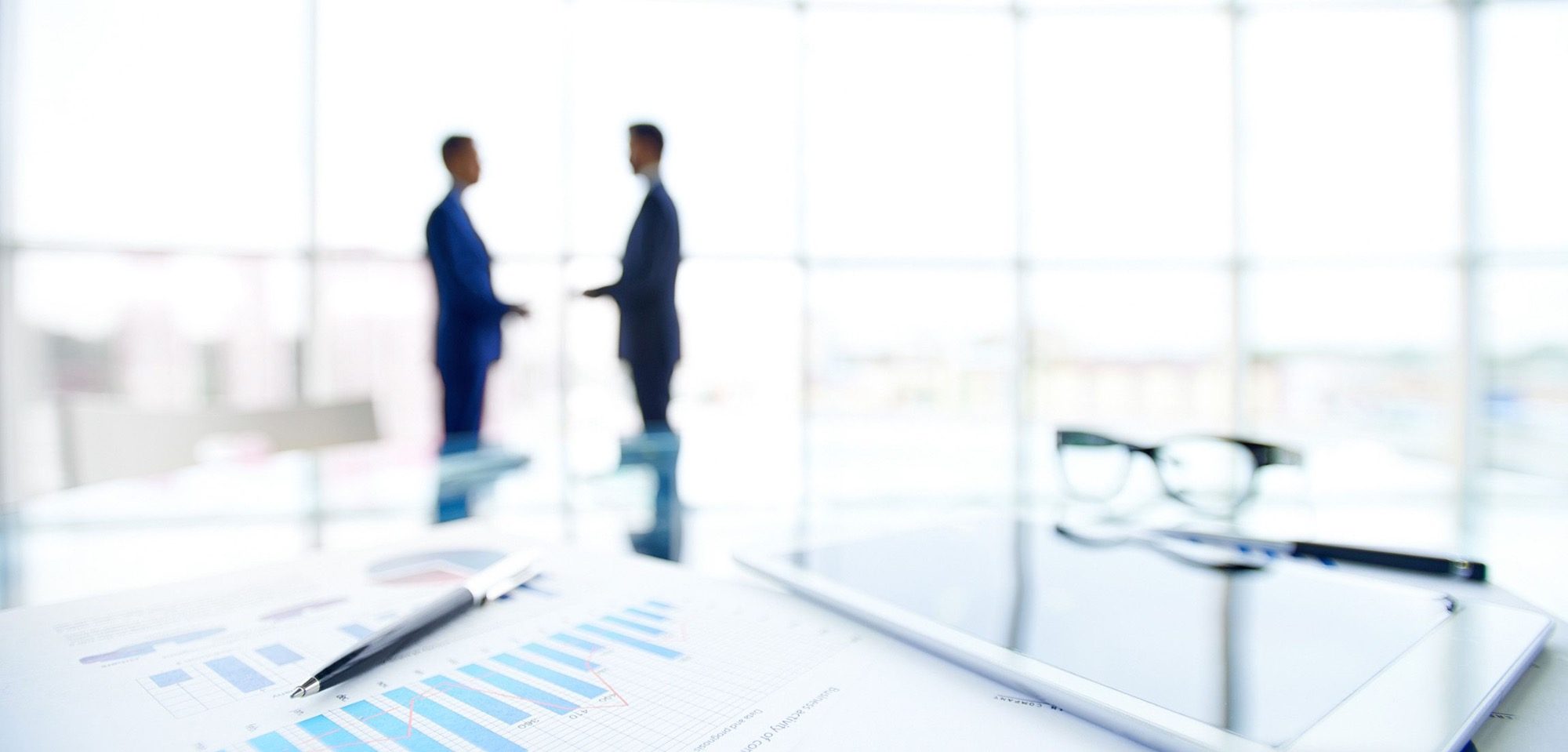 Begrüßung health & safety ehrl Zwei Männer in Anzügen stehen in einem modernen, hellen Büro vor einer großen Fensterfront mit Blick auf die Stadt.